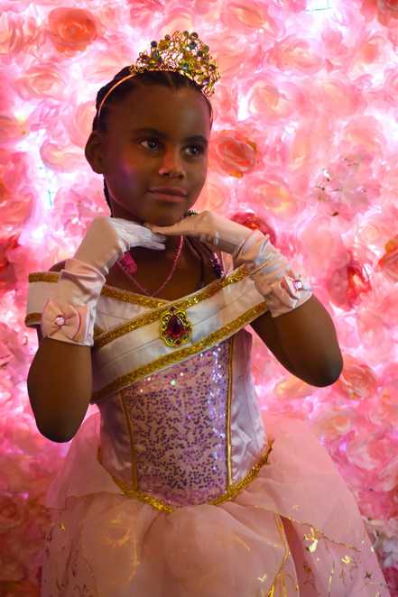 Avia poses in front of a wall of pink flowers wearing her pink princess outfit, resting both hands under her chin